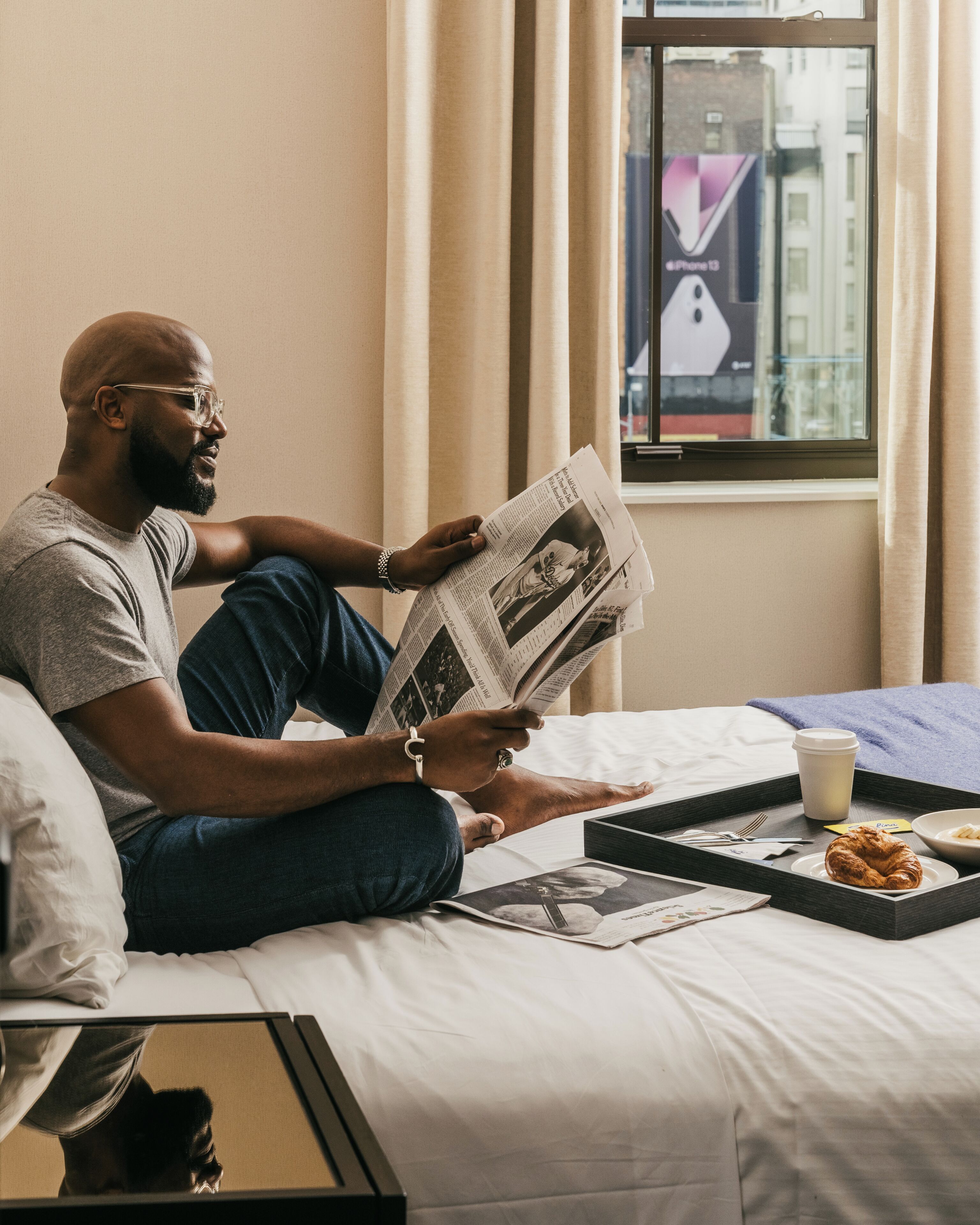 A person sitting on a bed reading a newspaper with a tray of food and a cup beside them.