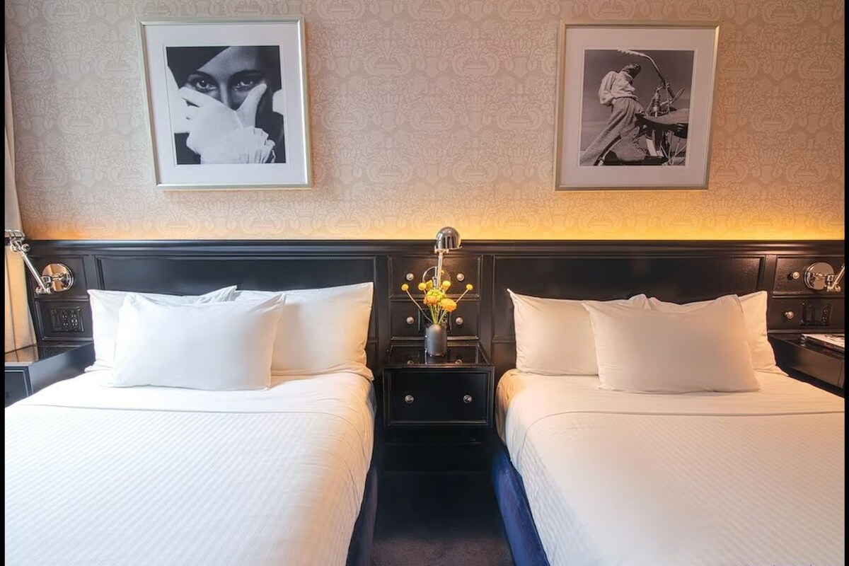 A hotel room with two beds, a nightstand with a lamp, and framed artwork on the wall.