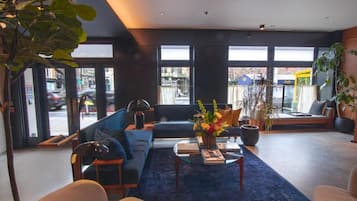 A modern hotel lobby with a blue sofa, a coffee table with books, and a large window offering a view of the street.