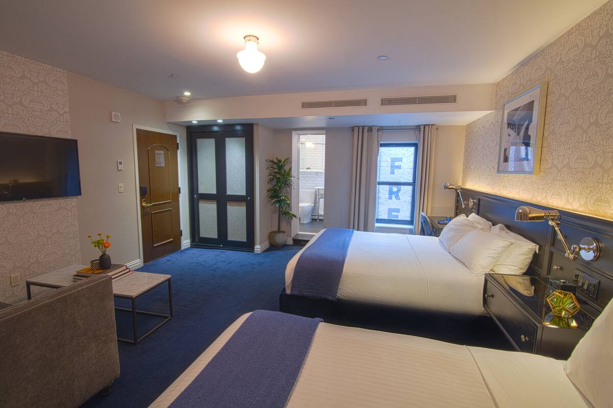 A hotel room with two beds, a desk, a TV, and a bathroom visible through an open door.