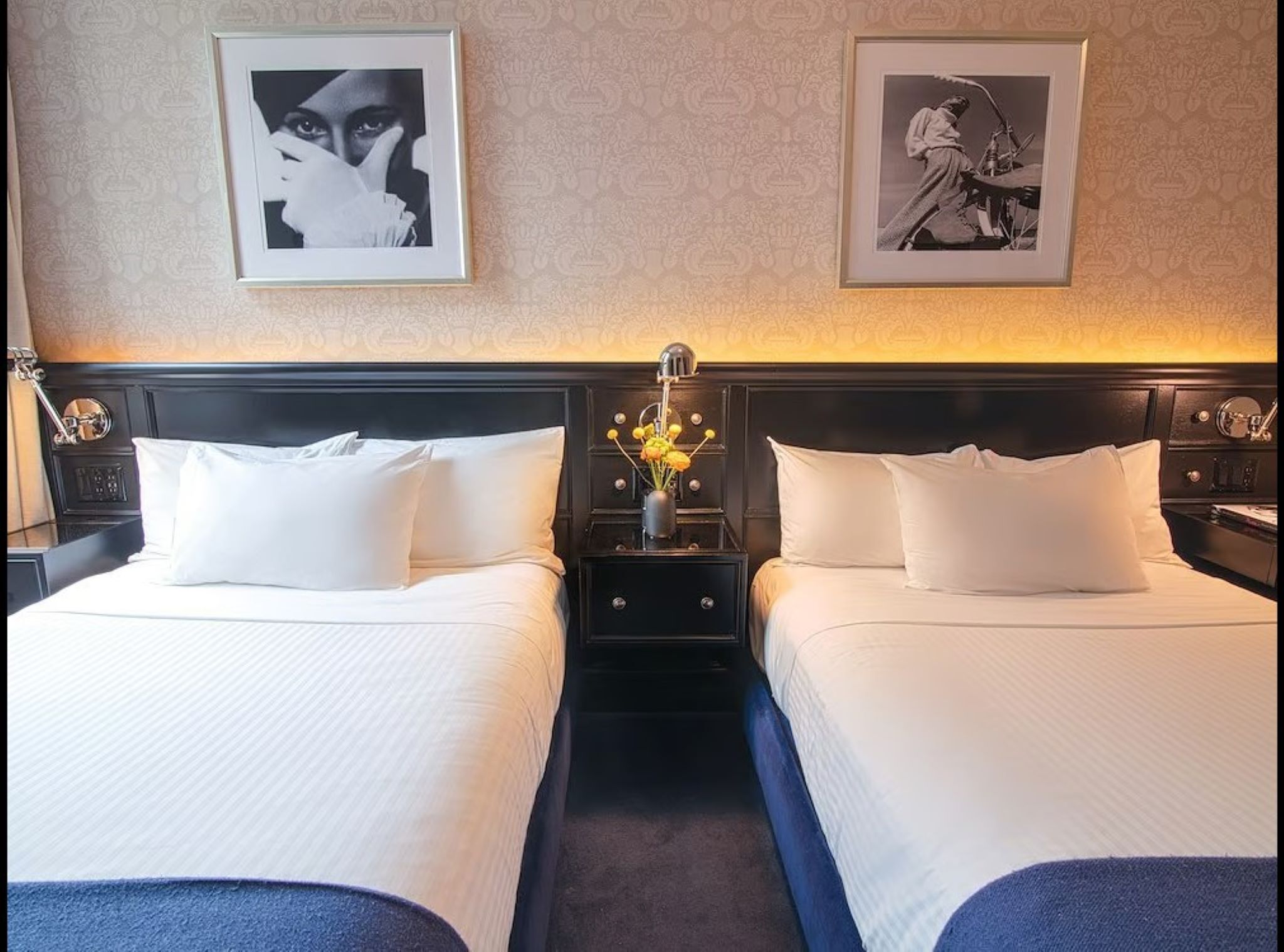 A hotel room with two beds, a nightstand with a lamp, and framed artwork on the wall.