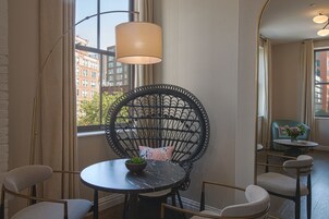A room with a round table, a black wicker chair with a cushion, and a view of buildings outside.