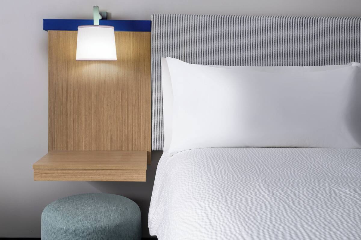 A neatly arranged hotel room with a wooden headboard, a bedside table with a lamp, a large bed with white bedding, and a small ottoman.