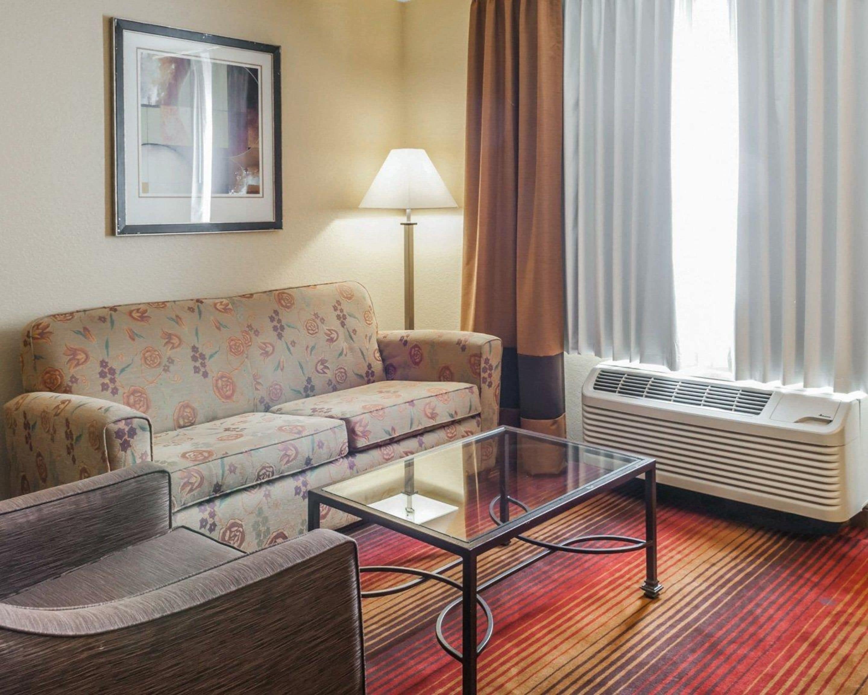 A hotel room with a floral sofa, a glass coffee table, a standing lamp, and a window with curtains.