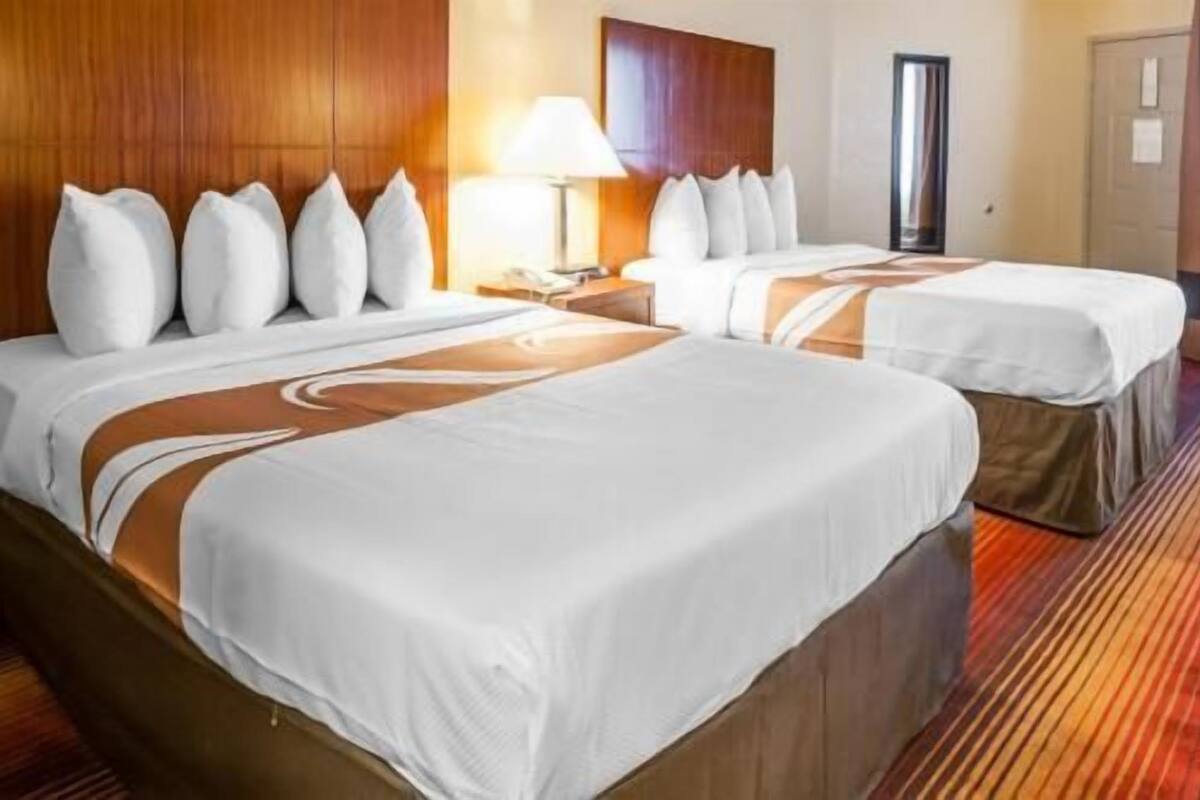 Two beds with white linens and brown accents, a wooden headboard, a nightstand with a lamp, and a door in the background.