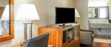 Habitación de hotel con escritorio de madera, dos lámparas, televisor de pantalla plana, minibar, microondas, sofá y baño con lavabo y espejo.