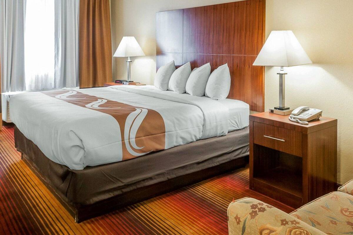 A hotel room with a large bed, two bedside lamps, a wooden headboard, a bedside table with a phone, and a patterned armchair.