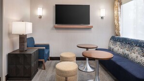 A modern hotel room with a flat-screen TV, a sofa, two ottomans, a lamp, and two small tables. - Hampton Inn Boulder - Louisville (Louisville)