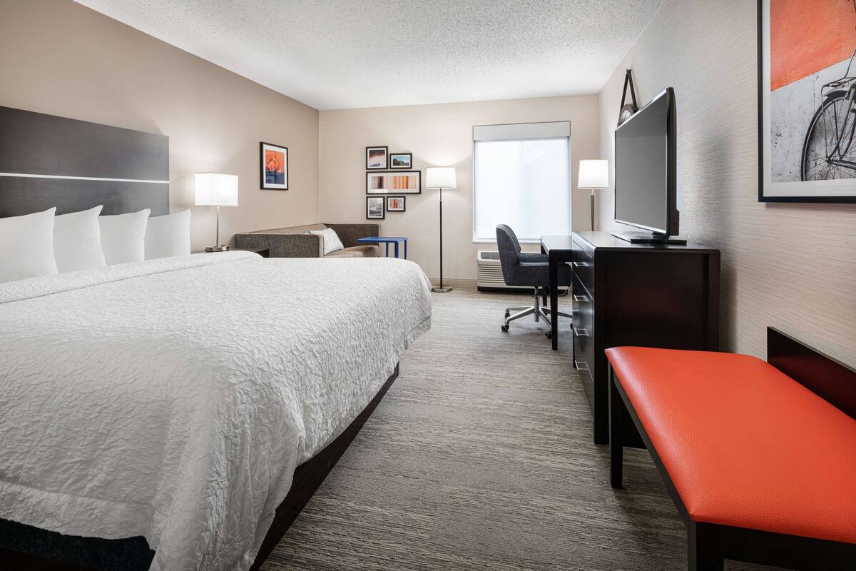 A hotel room with a large bed, a desk with a chair, a television, and a bench.