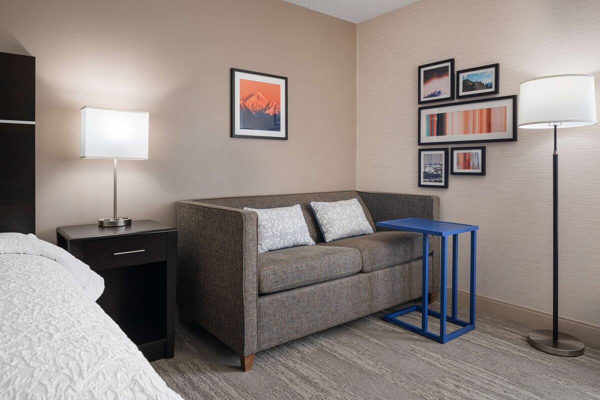 A hotel room with a grey sofa, a small blue side table, a black nightstand with a lamp, and a framed picture on the wall.