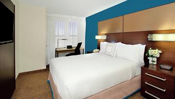 A hotel room with a large bed, a desk with a chair, a TV, and a nightstand with a lamp and flowers.