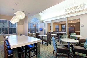 A hotel dining area with a large rectangular table, multiple booths, and round tables. There are chairs with patterned upholstery. The room has large windows and a ceiling light fixture. - Residence Inn by Marriott Colorado Springs South (Colorado Springs)