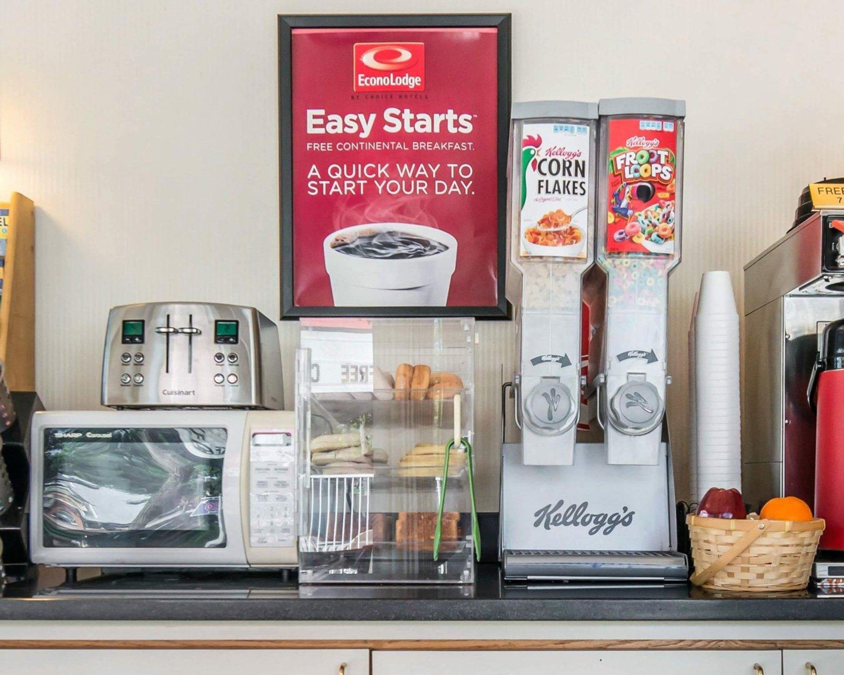 Una barra de desayuno con un tostador Cuisinart, un microondas, un refrigerador con pastelería y dos dispensadores de bebidas Kellogg's.