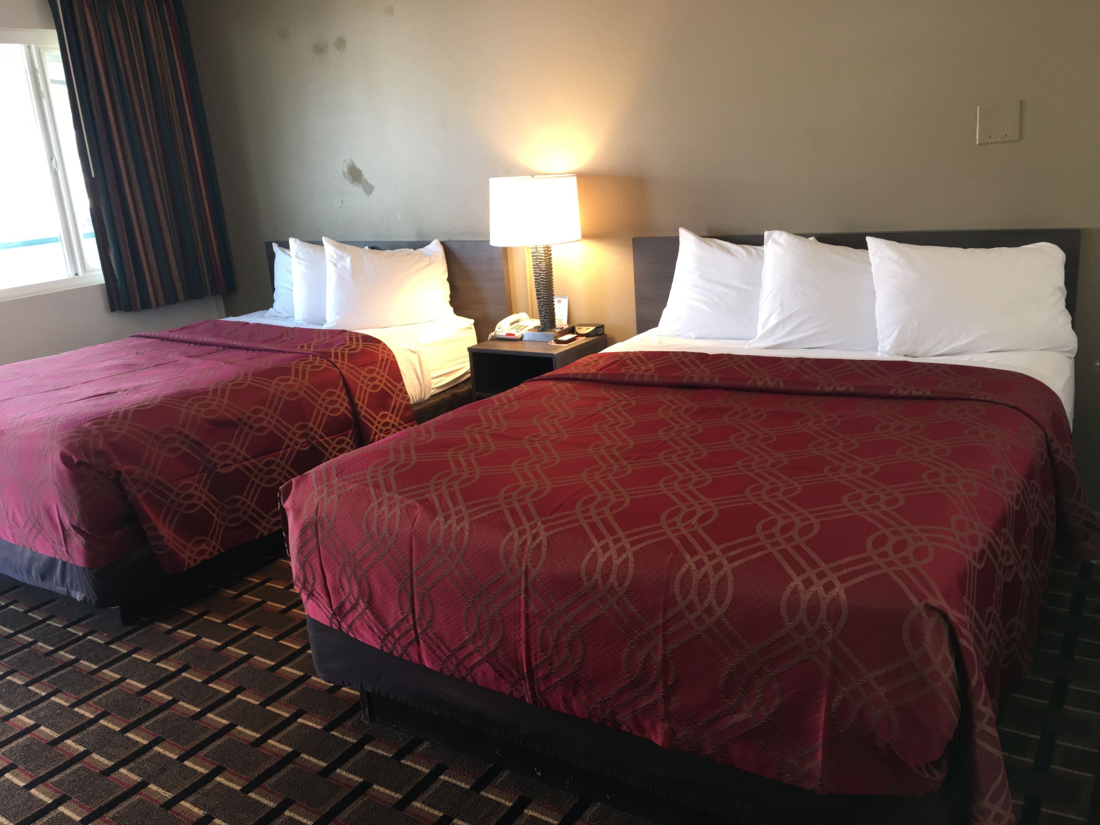Une chambre d’hôtel avec deux lits, chacun recouvert d’une couvre-lit à motifs rouges et agrémenté d’oreillers blancs. Une table de chevet avec une lampe et un téléphone se trouve entre les deux lits. La chambre est dotée d’un sol recouvert de moquette et d’une fenêtre avec des rideaux.