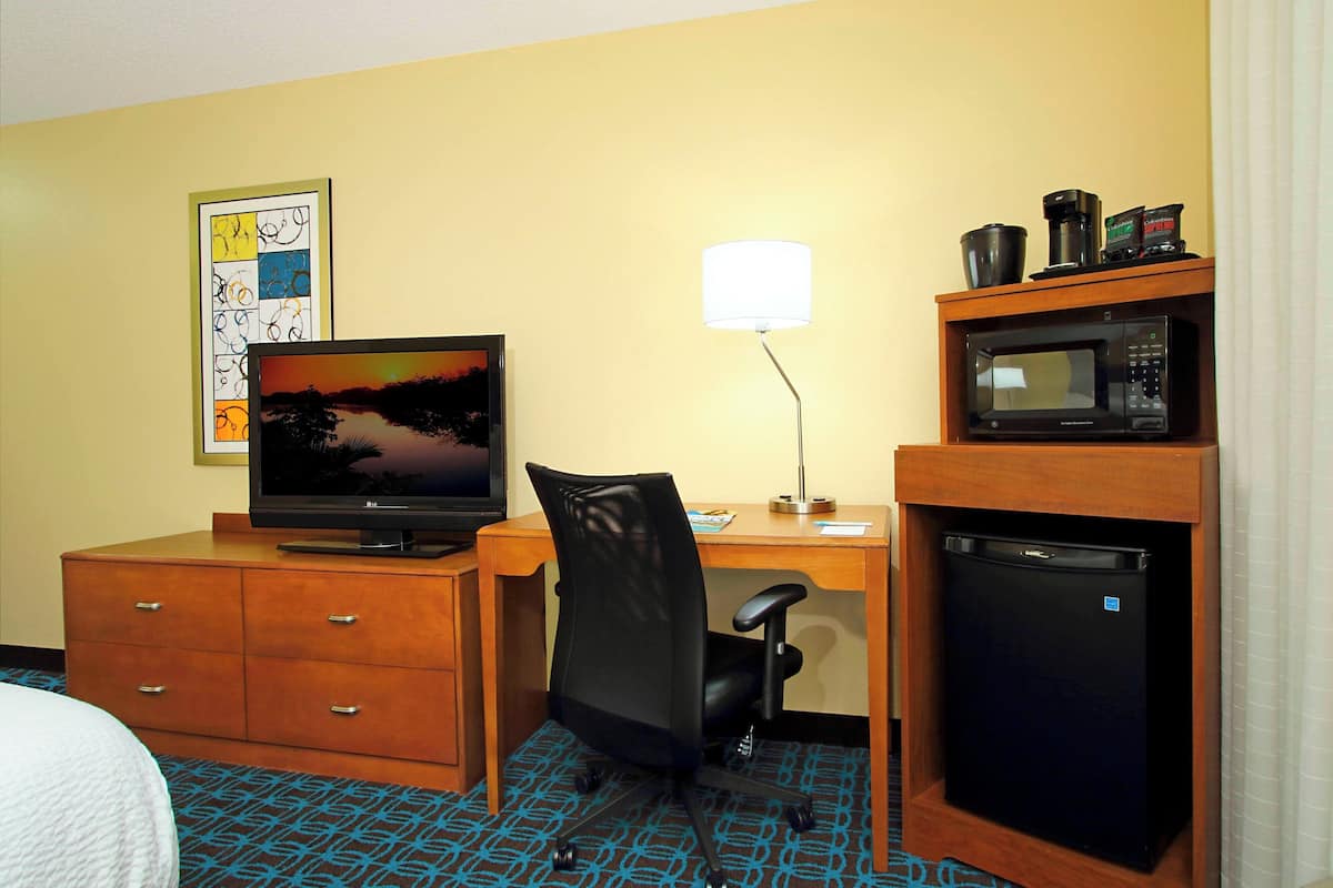 A hotel room with a desk, a computer, a chair, a microwave, and a refrigerator.