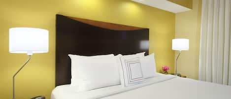 A neatly made bed with white linens, two bedside lamps, a digital clock, and a small table with a notepad and pen.