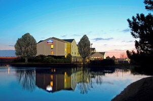 Un hotel con un letrero que indica que es un Fairfield Inn. Se encuentra junto a una masa de agua tranquila, con reflejos del edificio y los árboles.