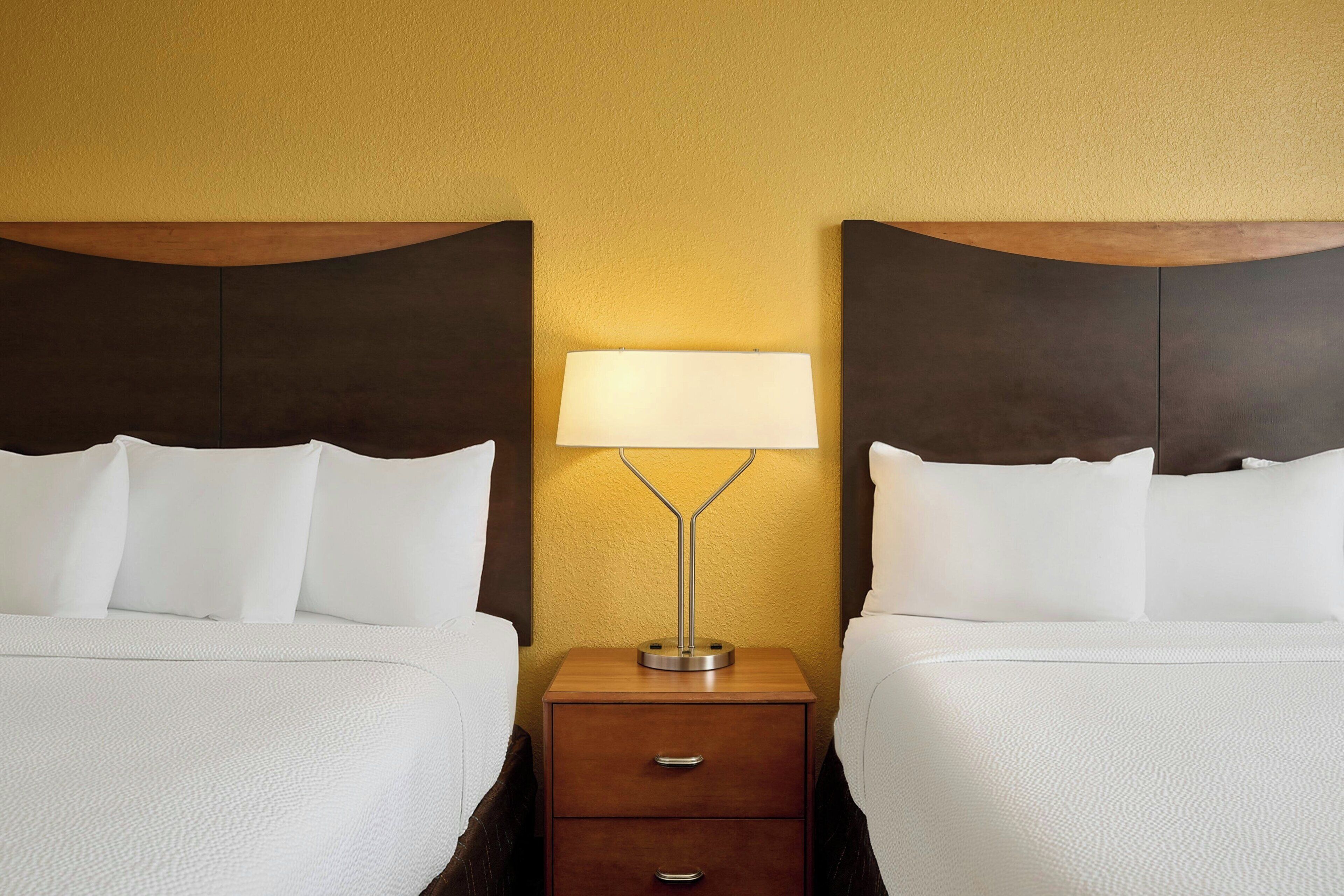 A hotel room with two beds, a nightstand with a lamp, and a wooden headboard.