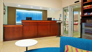 A hotel reception area with a wooden reception desk, a blue sofa with a colorful pillow, a small white table, and a vending machine. - Fairfield Inn & Suites by Marriott Colorado Springs South (Colorado Springs)