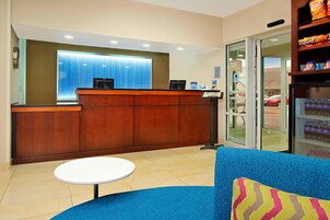 Zona de recepción de hotel con un mostrador de madera, un sofá azul con un cojín colorido, una pequeña mesa blanca y una máquina expendedora.