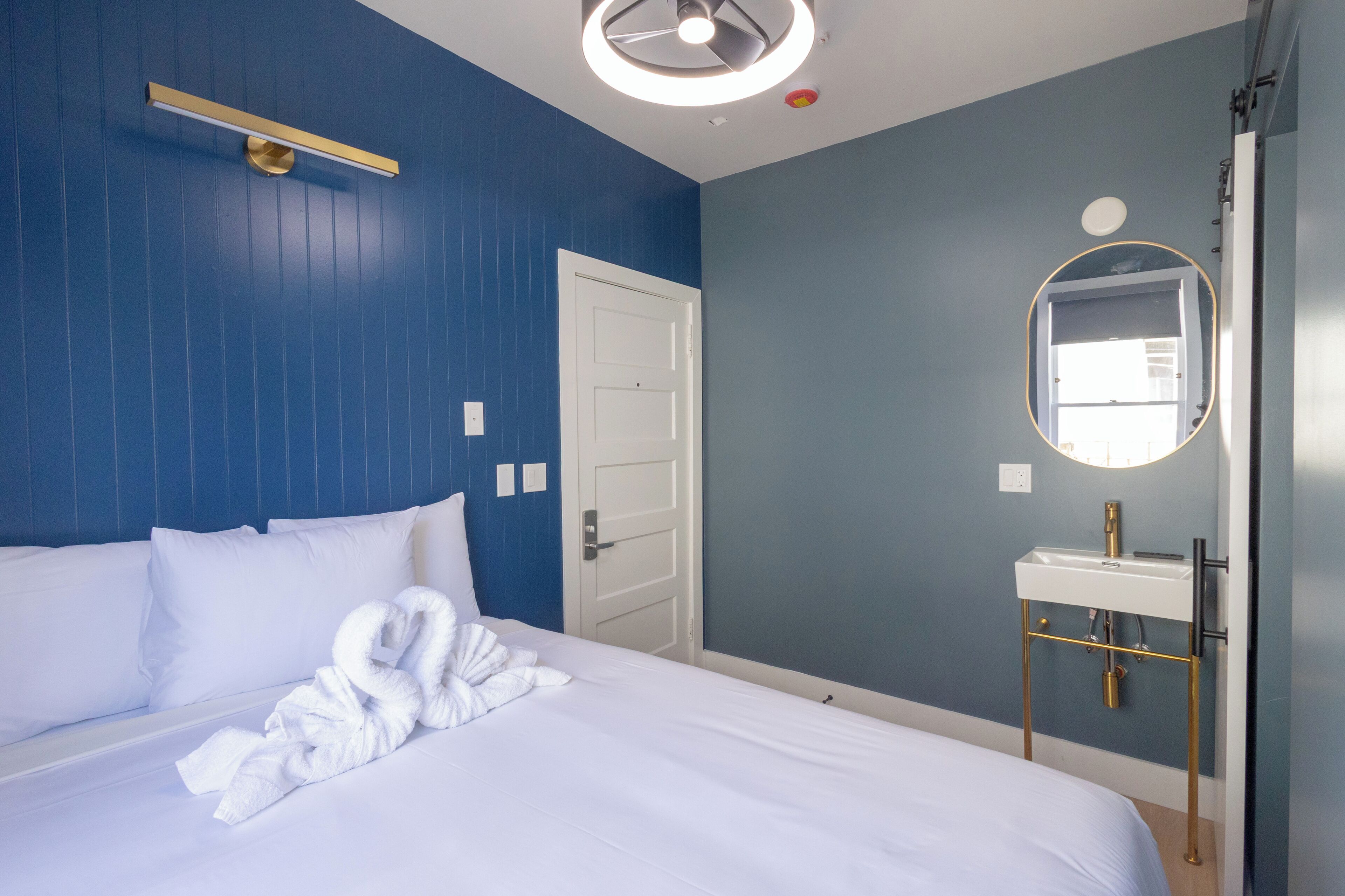 A hotel room with a bed, a white towel, a round mirror, a white sink, and a door.