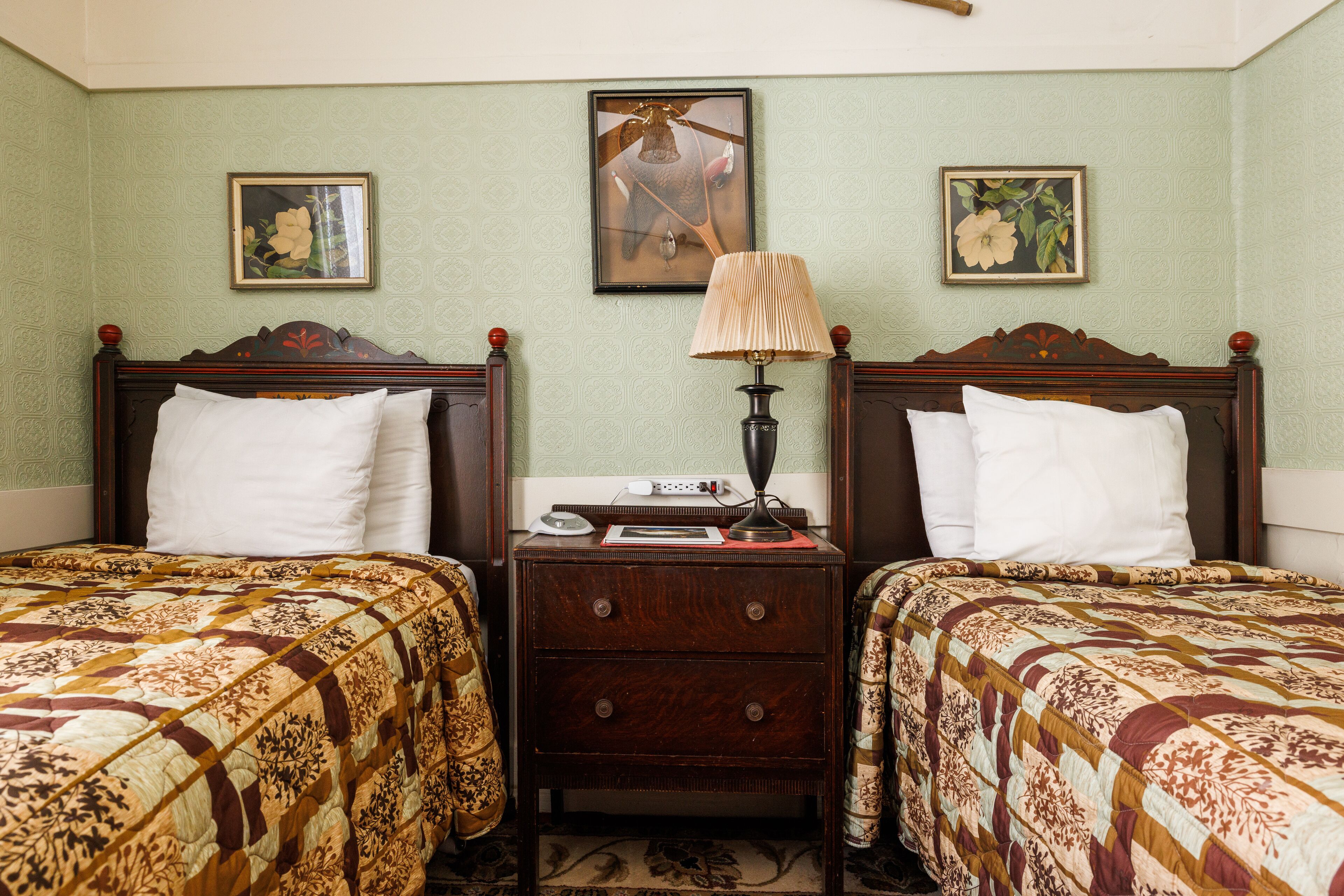 A room with two beds, each with a patterned bedspread, a nightstand with a lamp, and framed botanical prints on the wall.