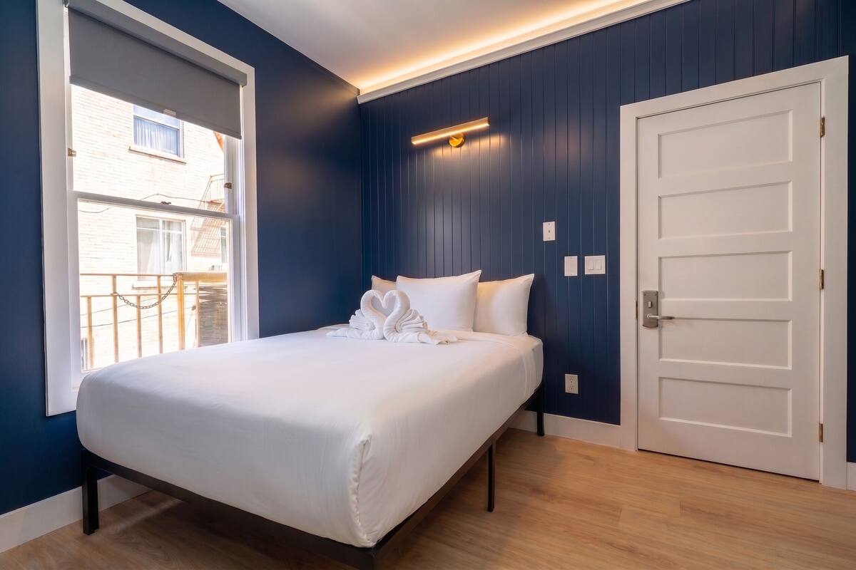 Un dormitorio con cama, una puerta blanca y ventana con vista a un edificio.