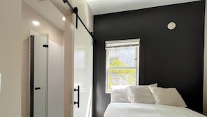 A modern bedroom with a bed, a window with blinds, a door, and a shower area. - San Remo Hotel (San Francisco)