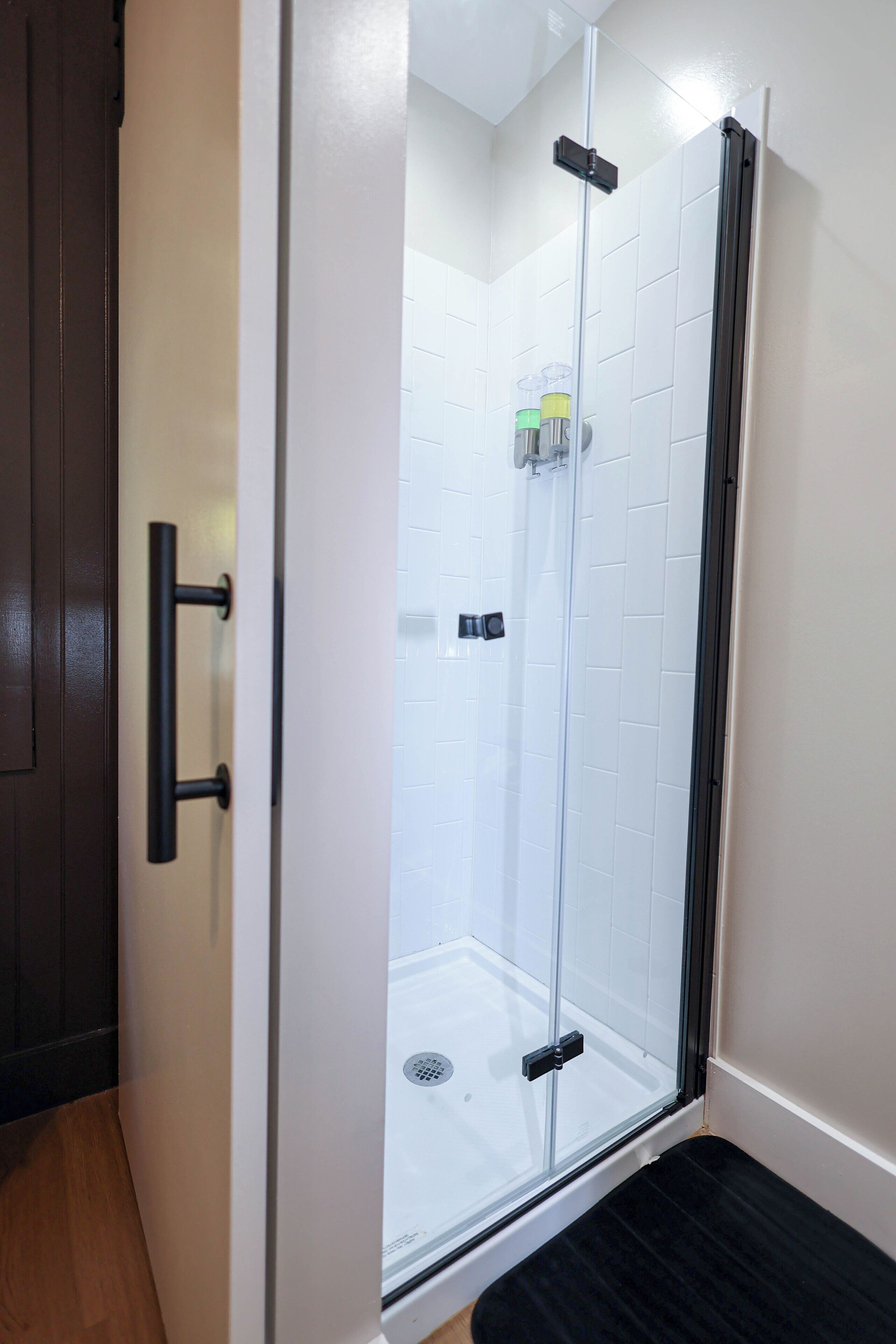 A glass-enclosed shower with a black door handle, white tiled walls, and a wooden floor.