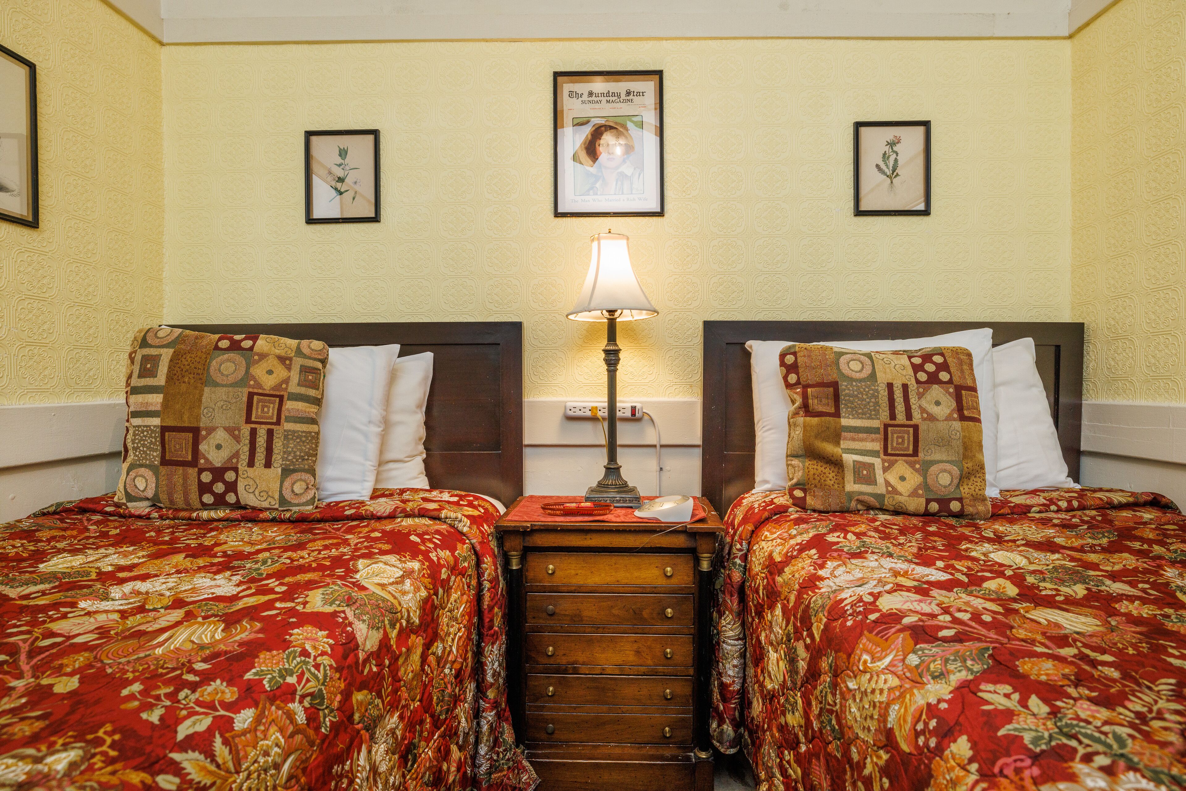 Una habitación de hotel con dos camas, cada una con una colcha de estampado floral y un cabecero de madera. Hay una mesita de noche entre las camas con una lámpara y un cuadro enmarcado en la pared de arriba.