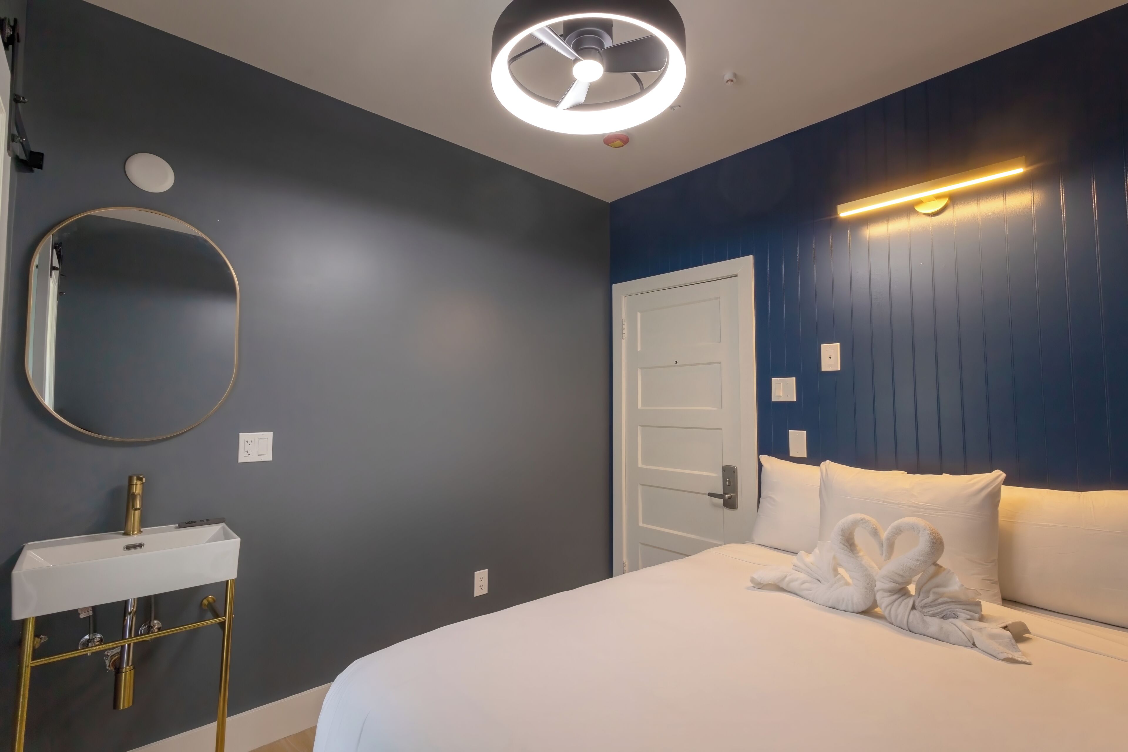 A hotel room with a bed, a round mirror, a white sink, and a door.