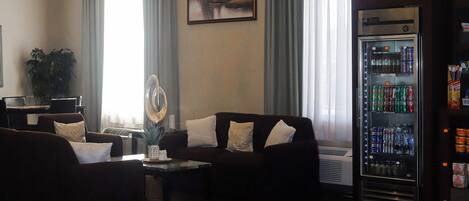 Una sala de estar moderna con un conjunto de sofás, una mesa de centro, una lámpara decorativa, un ventanal con cortinas y una máquina expendedora de bebidas.