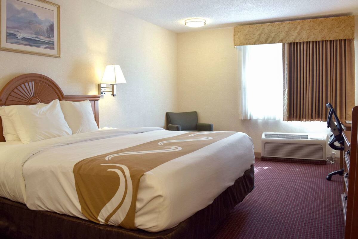 A hotel room with a large bed, a chair, an air conditioning unit, and a window with curtains.