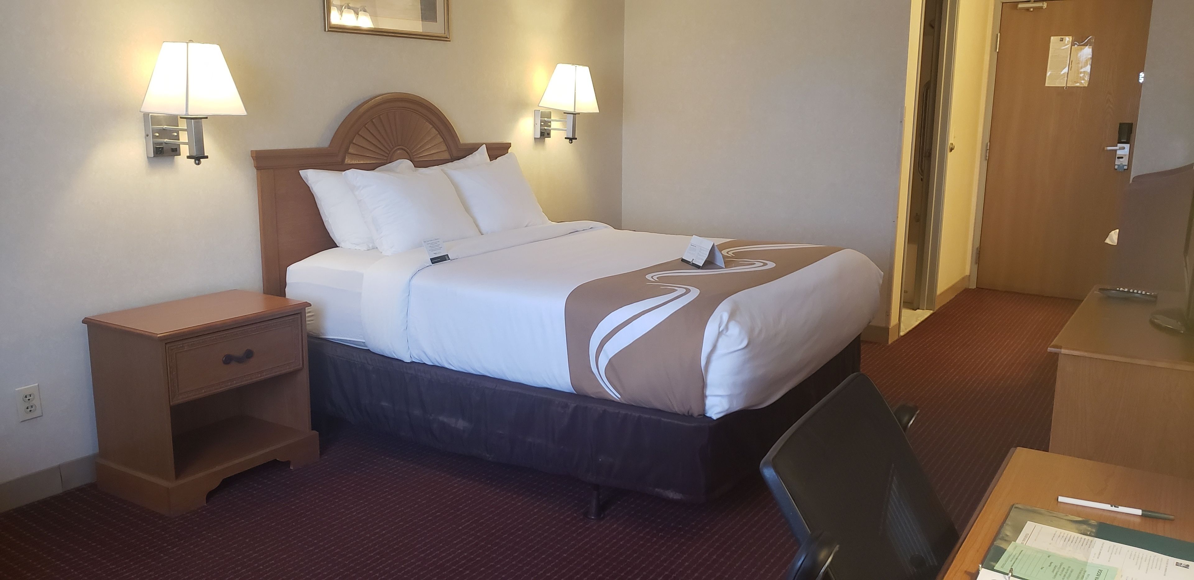 A hotel room with a bed, bedside lamps, a desk with a chair, and a door.