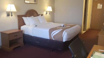 A hotel room with a bed, bedside lamps, a desk with a chair, and a door.
