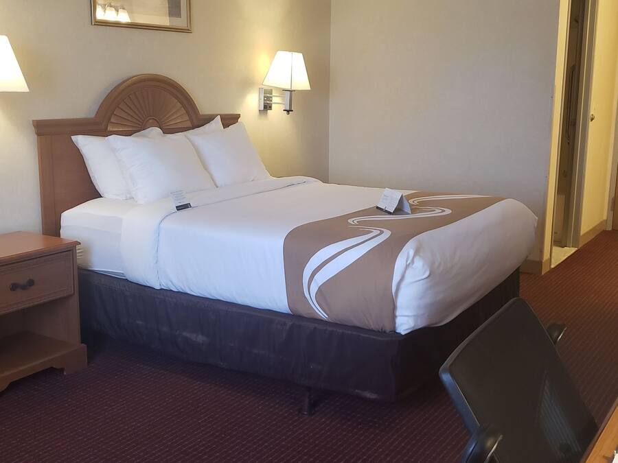 A hotel room with a bed, bedside lamps, a desk with a chair, and a door.