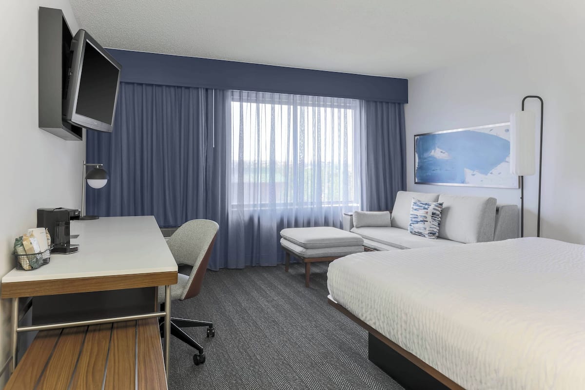 A hotel room with a bed, a desk with a chair, a television mounted on the wall, a coffee machine, and a window with curtains.