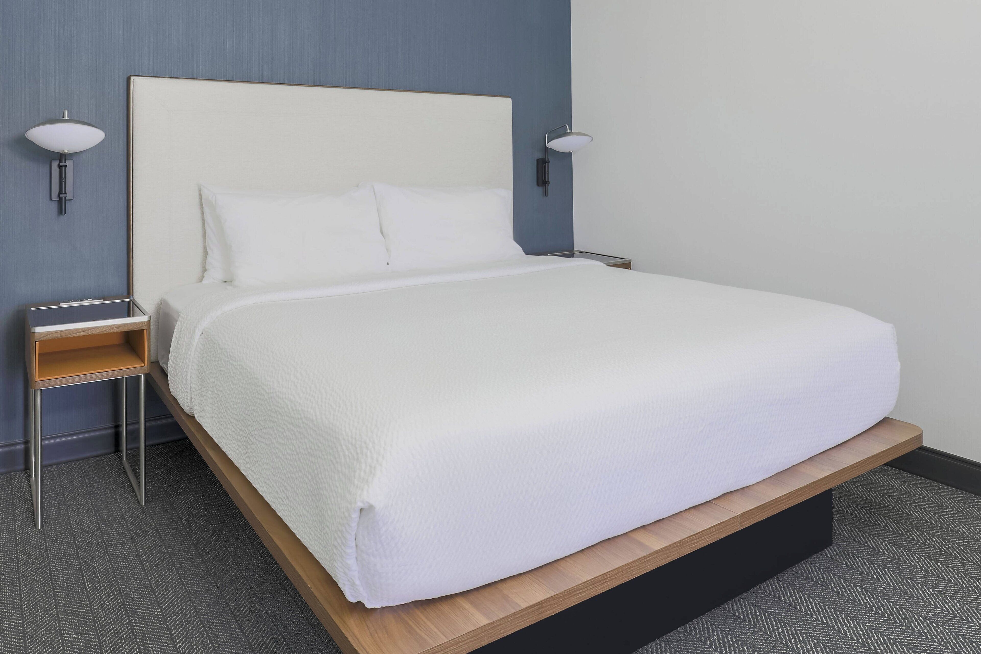 A neatly made bed with white bedding, a wooden platform, and a nightstand with a lamp.