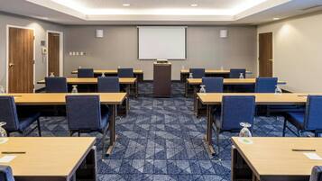 Un salón de conferencias con pantalla de proyector, varias mesas y sillas.