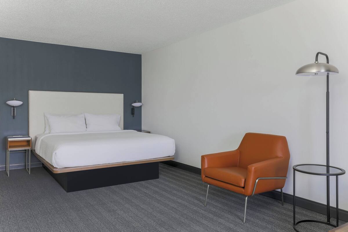 A hotel room with a bed, a chair, a lamp, and a small table.
