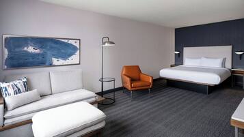 A hotel room with a bed, a sofa, an orange chair, a floor lamp, and a painting on the wall.