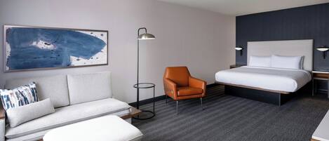 A hotel room with a bed, a sofa, an orange chair, a floor lamp, and a painting on the wall.