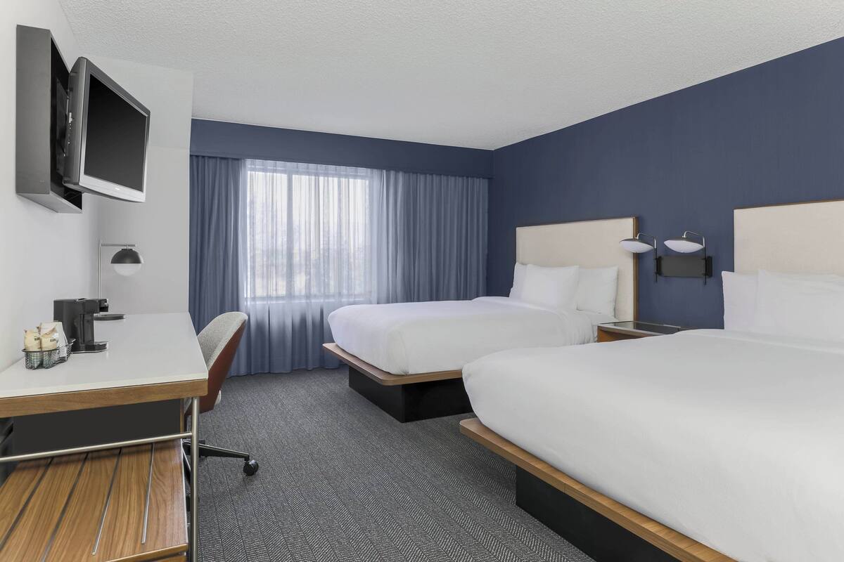 A hotel room with two beds, a desk, a TV, and a window with curtains.
