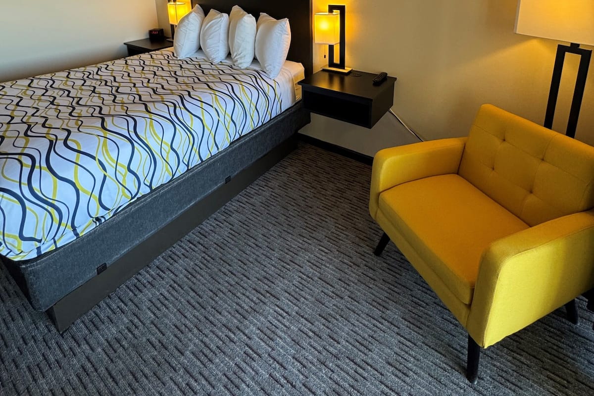 A hotel room with a bed, a yellow armchair, a nightstand with a lamp, and a wall decoration.