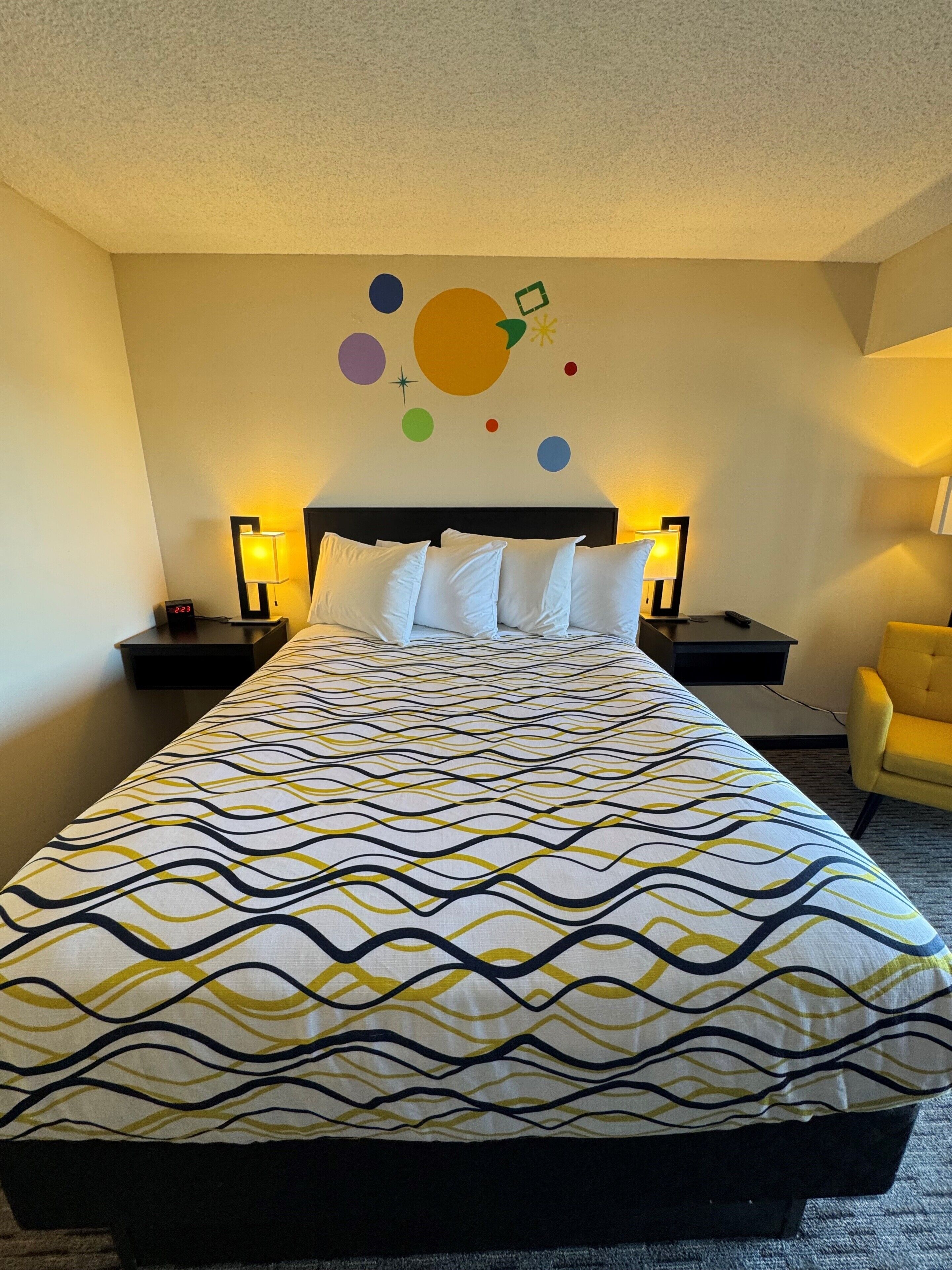 A hotel room with a bed, two bedside tables with lamps, a yellow armchair, and a wall with colorful circular decorations.