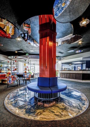A lobby with a central red and blue pillar, a circular seating area, and a glass table reflecting the room's interior. - Satellite Hotel (Colorado Springs)