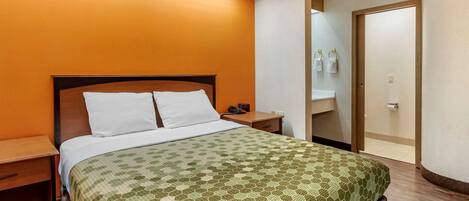 A hotel room with a bed, a nightstand, a bedside table, a bathroom with a sink and mirror, and a door.
