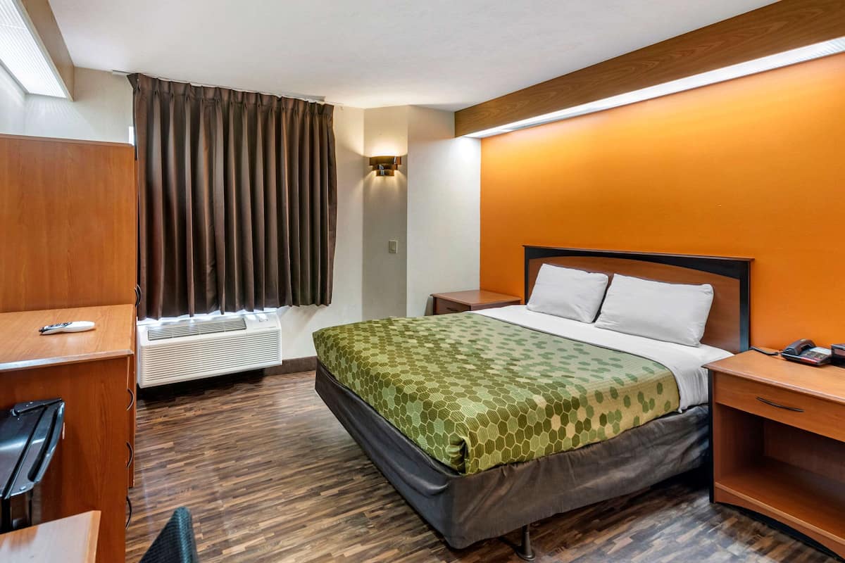 A hotel room with a bed, bedside table, desk, and an air conditioning unit.