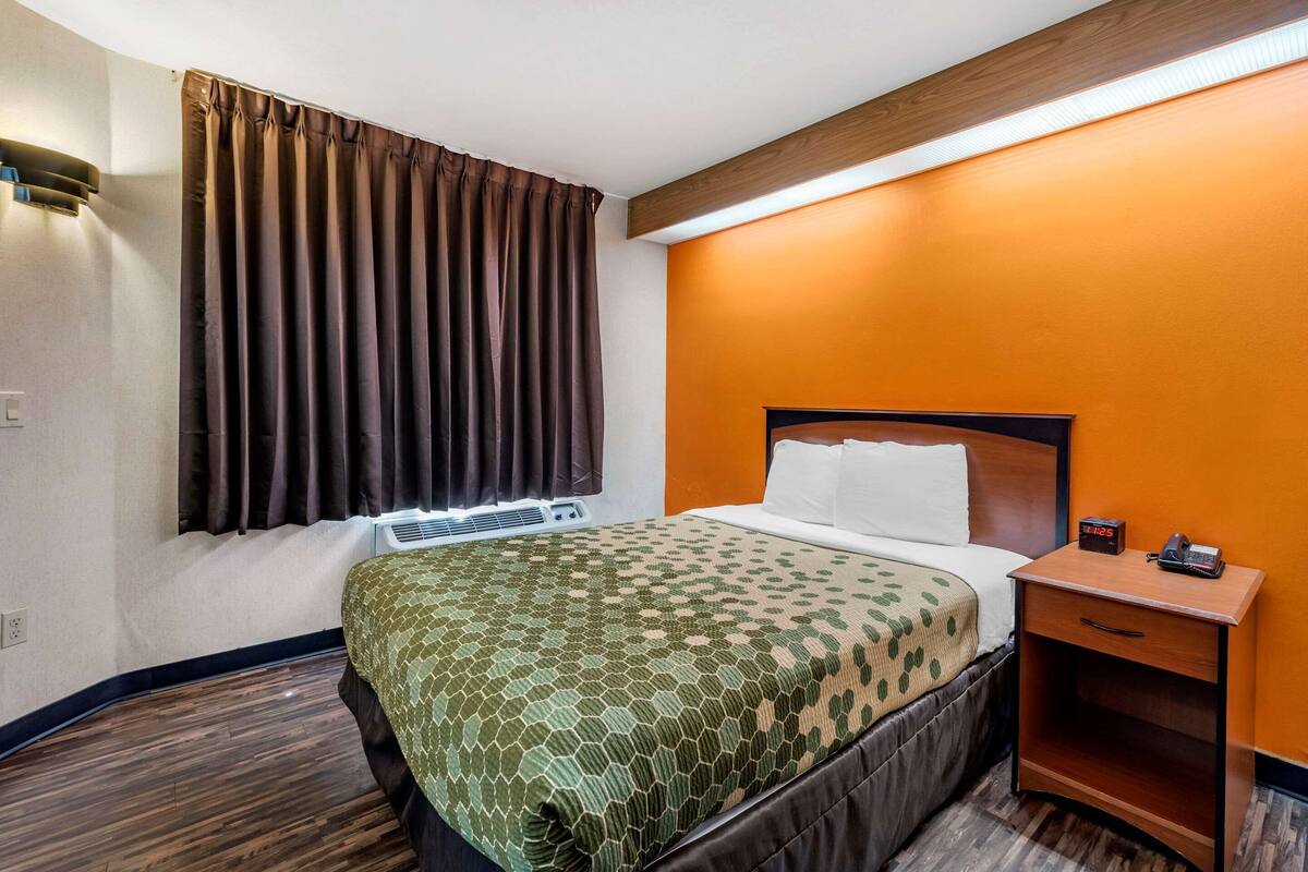 A hotel room with a bed, a nightstand, a heater, and a window with curtains.