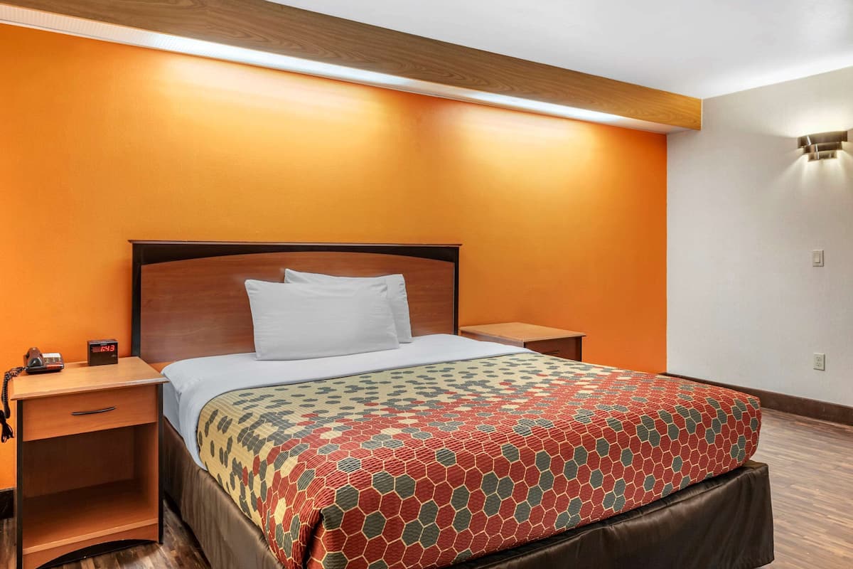 A hotel room with a wooden headboard, a bed with a patterned bedspread, a nightstand with a telephone, and a wall-mounted light fixture.